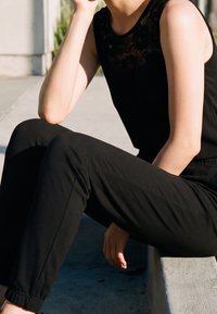 Personne assise dehors sur un rebord en béton portant un haut noir sans manches en dentelle et un pantalon noir, reposant le bras sur le genou à la lumière du soleil.