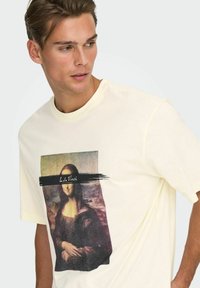 Kortærmet, lysegul T-shirt med trykt billede af Mona Lisa med en sort penselstrøg og tekstoverlæg. Blødt stof, afslappet pasform.