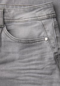 Primo piano di jeans in denim grigio chiaro che mostra la tasca anteriore, il passante per cintura e il dettaglio del rivetto metallico.
