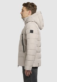 Svetlo bež pufasta jakna s kapuco, horizontalnim prešanjem, zadrgo na levem rokavu in prilegajočo silhueto. Črn logotip na rami.