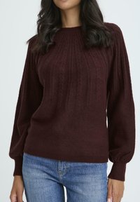 Maglione in maglia a treccia color bordeaux con collo rotondo a coste e maniche a palloncino. Texture morbida e vestibilità rilassata, abbinato a jeans blu.