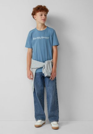 Hellblaues Baumwoll-T-Shirt mit weißem Schriftzug-Logo, kombiniert mit hellblauen weit geschnittenen Jeans und einem grauen Sweatshirt, das um die Taille gebunden ist. Weiße Turnschuhe.