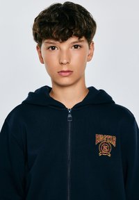 Mörkblå zip-up hoodie med en framficka med känguru-stil och en kontrasterande broderad logotyp i rött och gult på vänster bröst. Slät struktur.