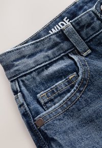 Traperice sa širokim pojasom, tamnoplave boje, vidljivim šavovima i malim prednjim džepom. Tekstura je blago izblijedjela i istrošena.