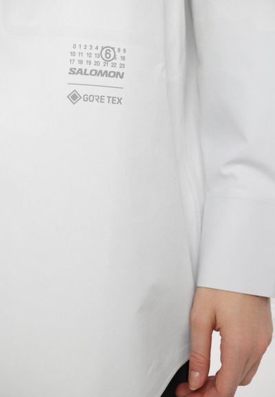 Balta nepralaidi marškiniai su Salomon logotipu, Gore-Tex etikete ir spausdintomis kalendoriaus grafikomis šonuose. Lygus tekstūra ir pritaikytas įsipareigojimas.