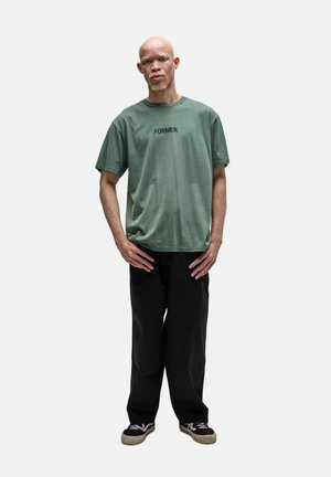 Homme grand avec albinisme portant un T-shirt vert marqué « FORMER », pantalon noir et baskets noires, debout les mains près des hanches.
