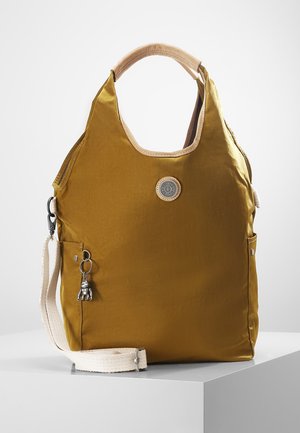 Musztardowożółta torba na ramię z beżowymi uchwytami i paskiem na ramię, z okrągłą naszywką z logo i małym srebrnym breloczkiem w kształcie małpki.