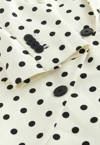 Tissu blanc à pois noirs avec une manche ornée de quatre boutons noirs et un bouton assorti à l'avant.