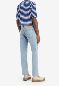 Muž v světle modrých rovně střižených džínách, béžových teniskách a modro-bílém pruhovaném tričku s krátkým rukávem, stojící zády k nám.