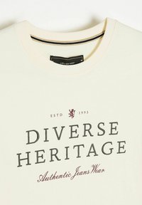 Jasna kremowa koszulka z bawełny, z okrągłym dekoltem. Nadrukowany tekst "Różnorodne Dziedzictwo" w czerni i winie, z graficznym wizerunkiem lwa.