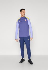 Sportslig træningsoverdel med en lilla top med gule detaljer og logo, parret med navy blå bukser. Fremstillet af åndbart stof.