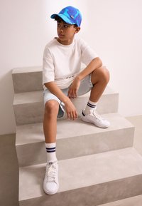 Mėlynas marmurinis kepuraitis, balti marškinėliai, šviesaus džinso šortai, balti sportbačiai ir dryžuotos kojinės. Sėdi ant šviesiai pilkų laiptų.