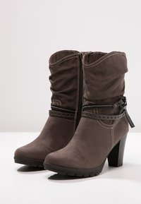 Botines de ante marrón con parte superior holgada, cremallera lateral, detalle de correa con tachuelas y tacón grueso. Suela de goma con acabado texturizado.