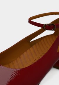 Κόκκινο παπούτσι με χαμηλό τακούνι από δερμάτινο πατέντα, με λουράκι στον αστράγαλο και μικρή χρυσή αγκράφα, με μαλακό ταμπά πέλμα με ανάγλυφες γραμμές.