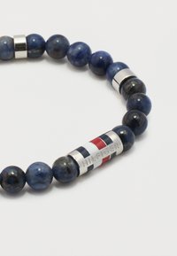 Ελαστικό βραχιόλι με στρογγυλές γυαλιστερές χάντρες σκούρου μπλε και μια ασημένια μεταλλική χάντρα με κόκκινες, λευκές και μπλε ρίγες και χαραγμένη τη λέξη "HILFIGER".