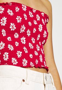 Červený top s nařasením a bílým potiskem sedmikrásky, nošený s bílo-světlými džíny s vysokým pasem, ukazující břicho a část ramene.