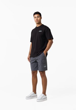 Man staat, draagt een zwart t-shirt, grijze cargoshorts met zijzak en witte sneakers voor een effen witte achtergrond.