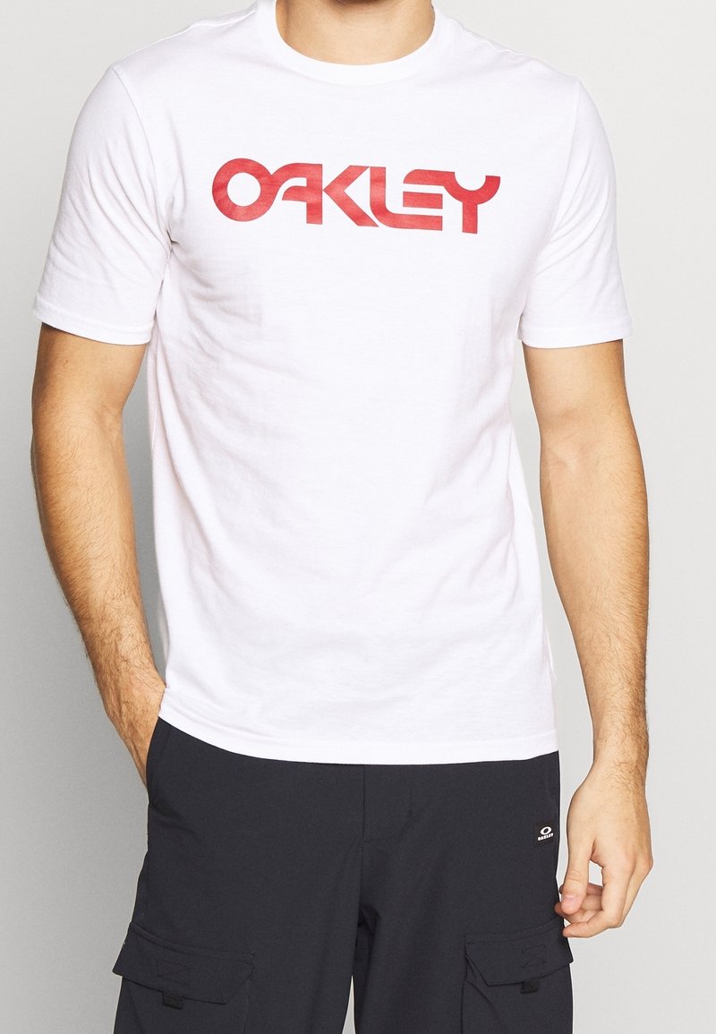 Bílé bavlněné tričko s výrazným červeným logem "OAKLEY" na hrudi. Krátké rukávy a standardní kulatý výstřih.