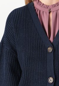 Gros plan sur un cardigan en tricot épais bleu marine avec de gros boutons, porté sur un chemisier mauve à col à volants et décolleté en trou de serrure.