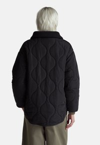 Černá prošívaná bunda s vlnitým vzorem, velkým límcem a texturovanou tkaninou. Volný střih s spadlými rameny a žebrovanými detaily.