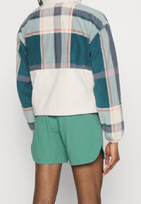 Kurtka polarowa z wzorem w turkusową kratę, beżową dolną częścią i elastycznymi mankietami, zestawiona z zielonymi szortami. Materiał o fakturze, luźny krój.
