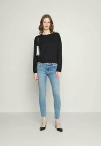 Svart langermet topp, lyseblå skinny jeans, sølvfarget skulderveske, svarte spisse leiligheter. Modellen står mot en nøytral bakgrunn.