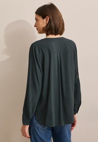 Blouse vert foncé à coupe ample, manches longues, poignets boutonnés et un subtil détail de plis à l'arrière. Tissu lisse et léger.