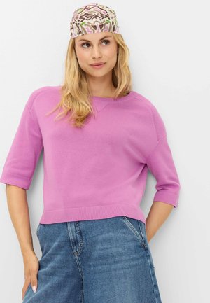 Kvinde med blondt hår iført et mønstret tørklæde, en lyserød top med korte ærmer og blå højtaljede jeans, står mod en hvid baggrund.
