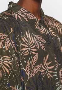 Hemd aus dunkelgrünem Stoff mit einem Blattmuster in Beige, Blau und Weiß. Verfügt über einen spitzen Kragen und ein lässiges Schnittdesign.