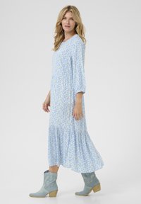 Gaiši zils ziedu apģērbs ar garām piedurknēm, savilktu jostu un viļņotu apakšmalu, kopā ar gaiši zilām kovboju zābakiem ar koka papēdi.