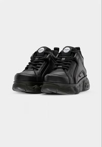 Mustad paksud paeltel tossud laineliste paksude taldadega ja brändiga keelekleepidega, esitletud ühevärvilise heleda tausta ees.