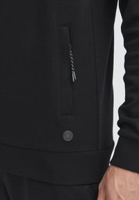 Sort sweatshirt med en lynlåslomme i siden, ribbede manchetter og kant. Har et rundt logo-patch nær kanten og tekstureret stof.