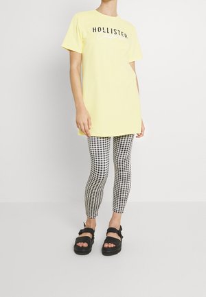 Person iført en gul Hollister t-shirt kjole, sort-hvide ternede leggings og sorte platformsandaler, stående på en hvid baggrund.