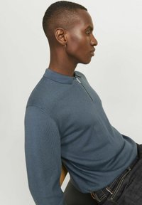 Langärmeliger Pullover mit Reißverschlusskragen aus dunklem blauen Strickstoff. Verfügt über einen klassischen Kragen und eine figurbetonte Silhouette, getragen mit dunklen Jeans.