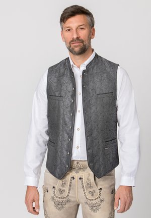 Mann trägt weißes Hemd, schwarze strukturierte Weste und beige bestickte traditionelle Lederhosen und steht vor schlichtem Hintergrund.