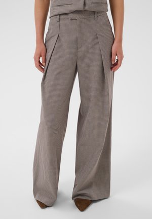 Personne portant un pantalon à carreaux taille haute et jambes larges avec des côtés plissés et des chaussures brunes pointues, debout devant un fond uni.