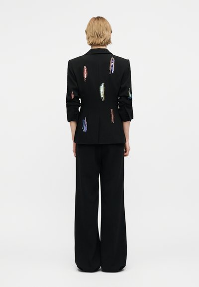 Άτομο με κοντά ξανθά μαλλιά φοράει μαύρο σακάκι και παντελόνι με φαρδιά πόδια· το σακάκι είναι διακοσμημένο με κατακόρυφες πολύχρωμες σειρές από παγιέτες.