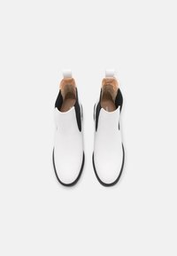 Bottines blanches avec des côtés élastiques noirs, dessus synthétique lisse, bout arrondi et intérieur contrastant beige. Équipées d'une languette à l'arrière pour un enfilage facile.