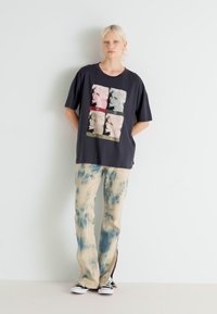 Camiseta gráfica negra con un diseño de collage multicolor, combinada con pantalones de tie-dye claro en tonos azul y beige y cremalleras laterales.