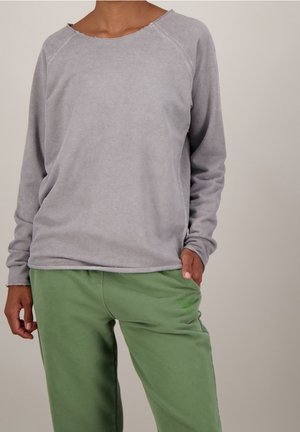 Person trägt ein hellgraues Langarmshirt und lockere grüne Hose, steht mit einer Hand in der Tasche vor einfarbigem Hintergrund.