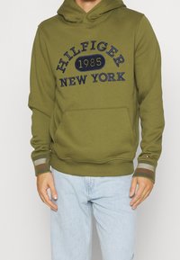 Sudadera verde oliva con un bolsillo canguro, que presenta el texto bordado en azul "HILFIGER 1985 NEW YORK." Puños acanalados con rayas en las muñecas.