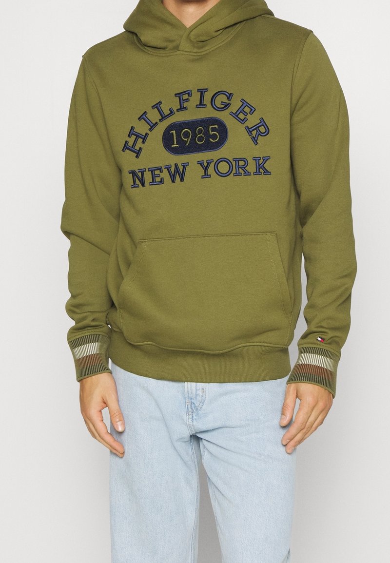 Sudadera verde oliva con un bolsillo canguro, que presenta el texto bordado en azul "HILFIGER 1985 NEW YORK." Puños acanalados con rayas en las muñecas.