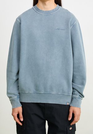 Personne portant un sweat-shirt bleu délavé à col rond avec un petit logo sur la poitrine et un pantalon cargo noir avec des logos en patch sur les poches.