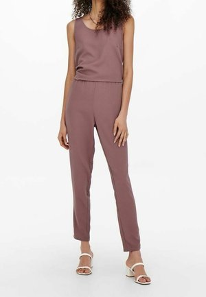 Vrouw draagt een mouwloze mauve jumpsuit met een elastische taille en witte sandalen met blokhak en open teen, staand tegen een witte achtergrond.