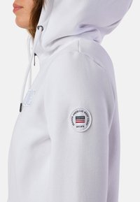 Weißes Kapuzensweatshirt mit Kordelzug und einem runden Patch am linken Ärmel, das Logo- und Textdetails zeigt. Weicher Stoff.