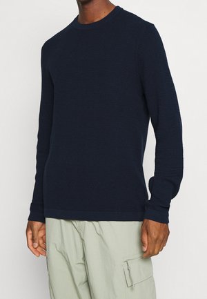 Dunkelblaues Langarm-Pullover mit Rundhalsausschnitt und strukturiertem Strickmuster, mit gerippten Bündchen und einem einfarbigen Saum. Über hellen Cargo-Hosen getragen.