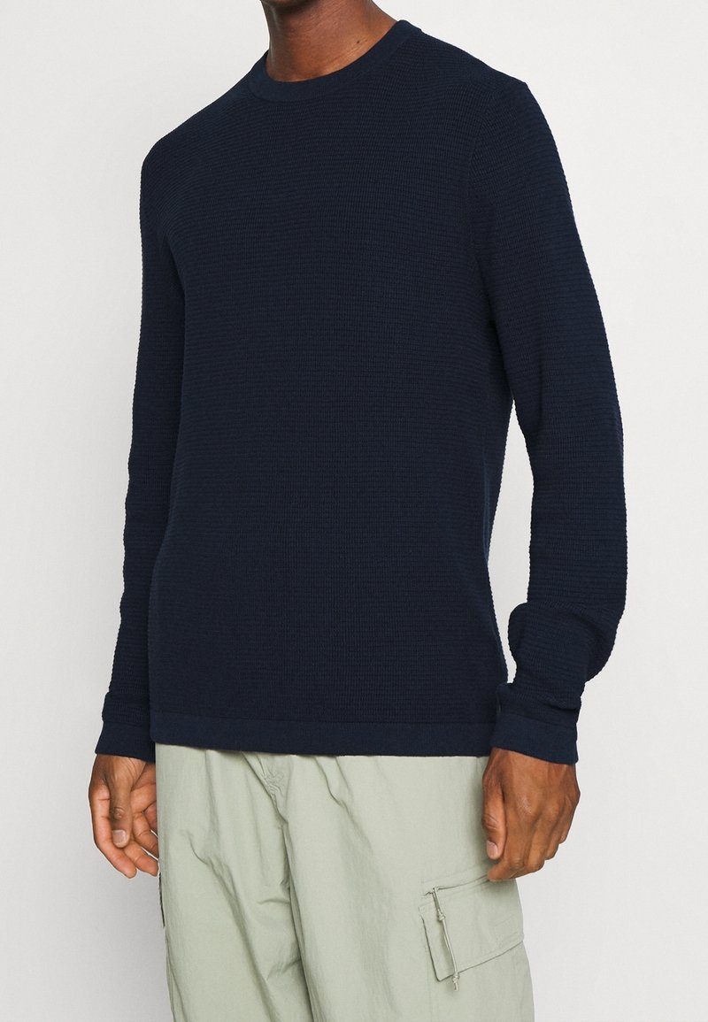 Suéter de cuello redondo de manga larga en azul marino con un patrón de punto texturizado, que presenta puños acanalados y un dobladillo sólido. Llevado sobre pantalones cargo claros.