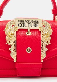 Borsa a mano in pelle rossa con dettagli decorativi in oro, dotata di una placca logo prominente e dettagli impressi. Design compatto e strutturato.