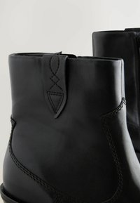 Botines de cuero negro con textura suave, que presentan costuras decorativas y un tabique triangular en el lateral para facilitar su uso.