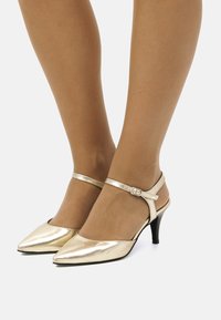 Zapatos de tacón alto metálicos dorados con punta afilada y correa en el tobillo, que presentan una suela negra y una textura suave.